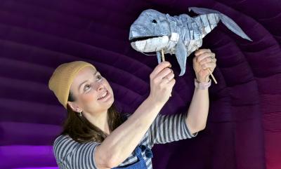 A woman in a hat and dungarees holding a whale puppet