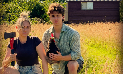A man holding a chicken in a field and a woman holding an axe